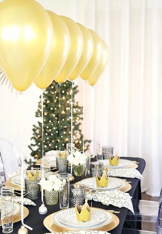 a shiny black and gold NYE party tablescape with a black tablecloth, gold chargers and gold balloons over the table, grey glasses and white blooms