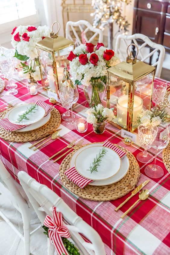 a stylish Christmas table with a red plaid tablecloth, gold lanterns, red and white blooms and woven placemats