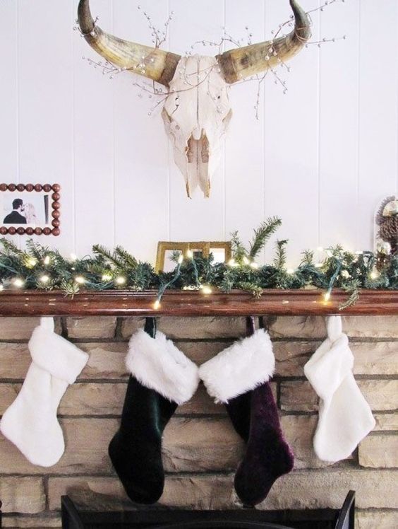 a stylish boho Christmas mantel with white, dark green and purple velvet stockings, evergreens and lights is cool