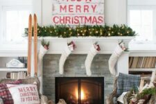 a traditional Christmas mantel with a light evergreen garland, white stockings, a sign, skis and some plaid touches here and there