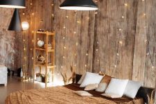 a warm-colored bedroom with wooden walls, a low bed with earthy-toned bedding, a wooden storage unit, a rattan stool and string lights covering the wall