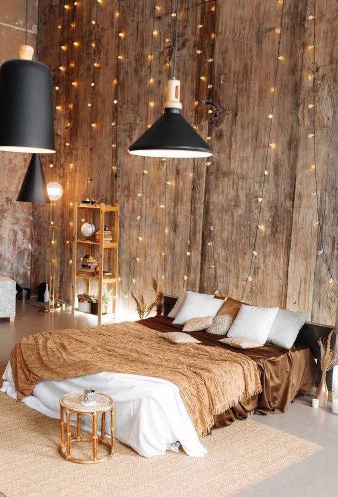 a warm colored bedroom with wooden walls, a low bed with earthy toned bedding, a wooden storage unit, a rattan stool and string lights covering the wall