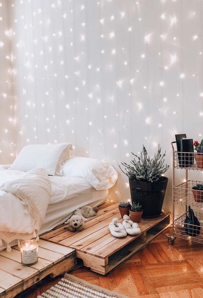 a welcoming bedroom with a pallet bed and white bedding, a metal shelving unit, potted plants and a whole curtain of string lights