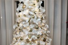a white Christmas tree with gold and white ornaments, white ruffle ribbons and a lit up tree topper