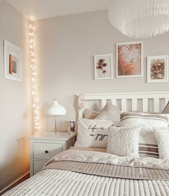 an airy and chic bedroom with a white bed and nightstands, neutral bedding, a gallery wall and a fringe chandelier, string lights hanging down from the ceiling