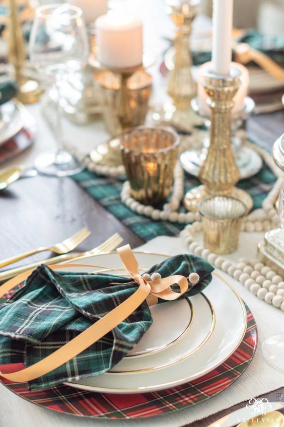 an elegant Christmas tablescape with red plaid chargers and green plain linens, wooden beads and mercury glass candleholders
