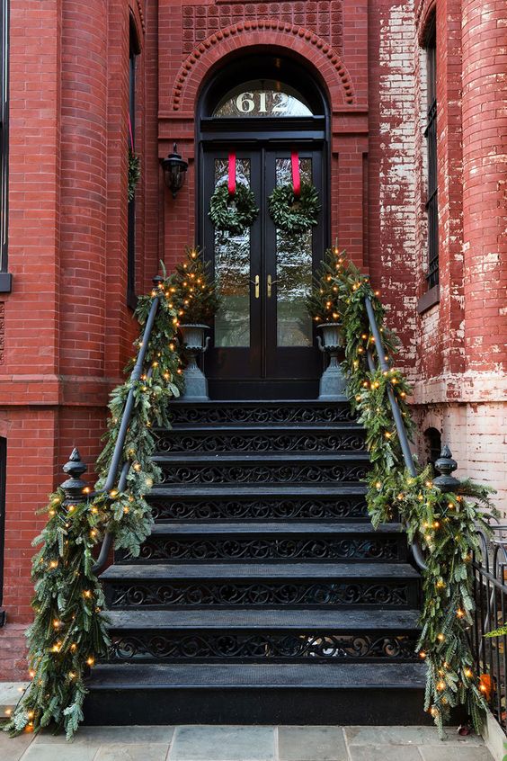 a beautiful festive porch with evergreen garlands and lights, mini Christmas trees and wreaths with red ribbons is amazing