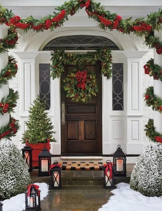 a bold and elegant Christmas porch with an evergreen lit up garland, a wreath with a red bow, a greenery and red ribbon garland, a mini tree, candle lanterns