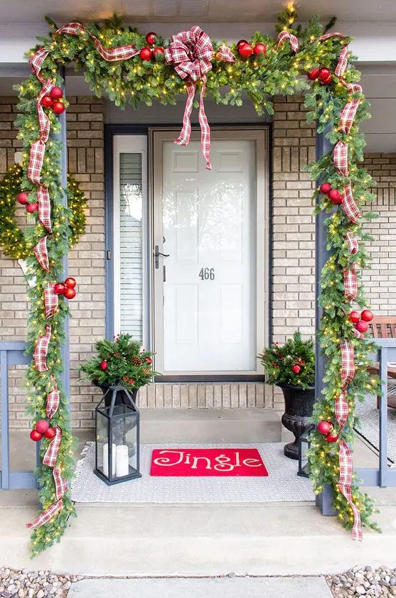 a bright Christmas porch with an evergreen garland with lights, red ornaments, plaid ribbons and bows, a red rug, candle lanterns and arrangements