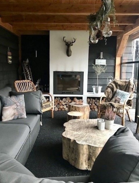 a covered winter terrace with a large rug, grey furniture, rattan chairs, a tree stump table, a built in fireplace and firewood is very cozy