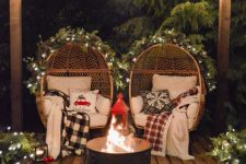 a cozy Christmas decor with egg-shaped chairs with lots of pillows and blankets, a hearth, a lantern, lights and urns with lights and evergreens