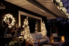 a fab Christmassy deck with a neutral sofa, a white side table, a pendant chair, Christmas trees and a wreath and candle lanterns