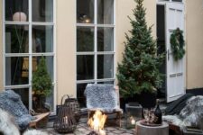 a gorgeous winter or Christmas terrace with wooden furniture, a cocnrete mini table, a fire bowl, candle lanterns, a Christmas tree with lights and faux fur
