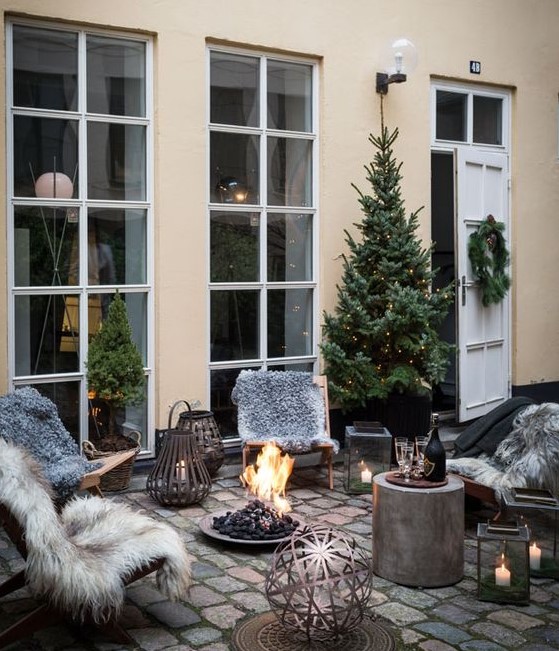 a gorgeous winter or Christmas terrace with wooden furniture, a cocnrete mini table, a fire bowl, candle lanterns, a Christmas tree with lights and faux fur