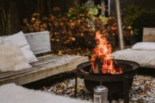 a lovely winter patio with pebbles on the ground, a metal fire bowl, wooden benches covered with faux fur and faux fur pillows