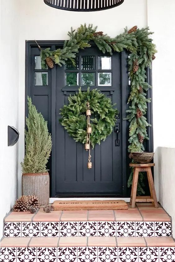a very natural Christmas porch with a tree, pinecones, an evergreen wreath with bells and an evergreen garland with pinecones