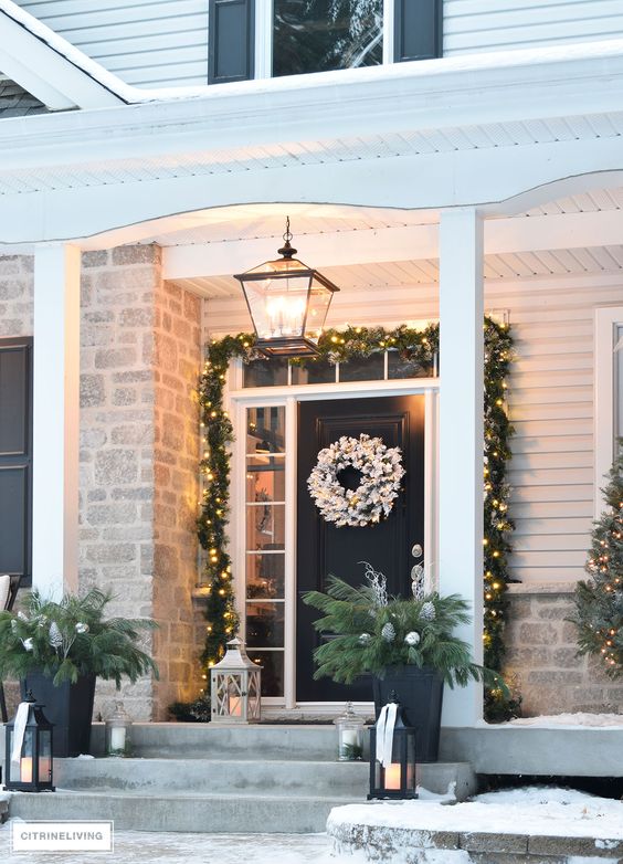 an evergreen and light garland framing the entrance door, a flocked evergreen wreath, candle lanterns and evergreen arrangements with pinecones