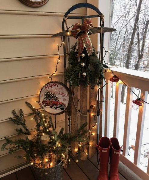 red rubber boots, some firewood and evergreens in a bucket and a vintage sleigh with bells and evergreens plus lights around