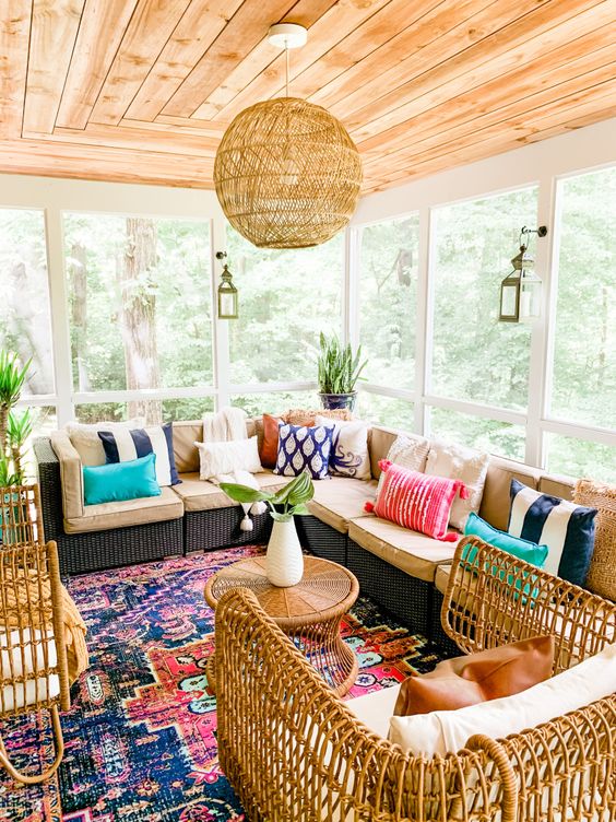 a colorful boho sunroom with a wicker sectional, colorful pillows and a rug, pendant lanterns and a lamp, wicker chairs