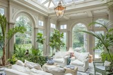 a lush sunroom or orangery with neutral furniture, tan poufs, a pendant lamp and lots of potted greneery and blooms