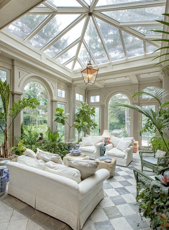 a lush sunroom or orangery with neutral furniture, tan poufs, a pendant lamp and lots of potted greneery and blooms