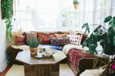a pretty and bright boho sunroom with a sectional, colorful pillows and blankets, a carved wooden table, a leather pouf and potted plants