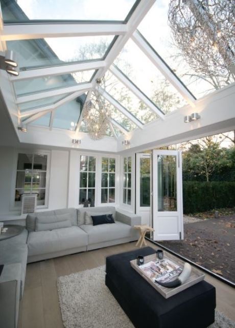 a simple and contemporary sunroom with a large grey sectional, a black ottoman, a vintage chandelier and folding doors