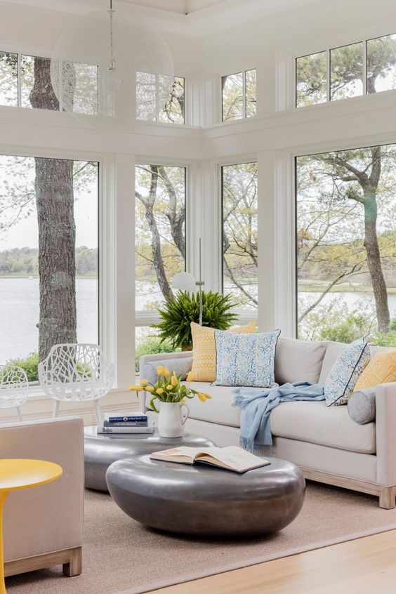 a beautiful modern sunroom with a neutral sofa, printed pillows, pebble-like tables, white chairs and potted plants plus views
