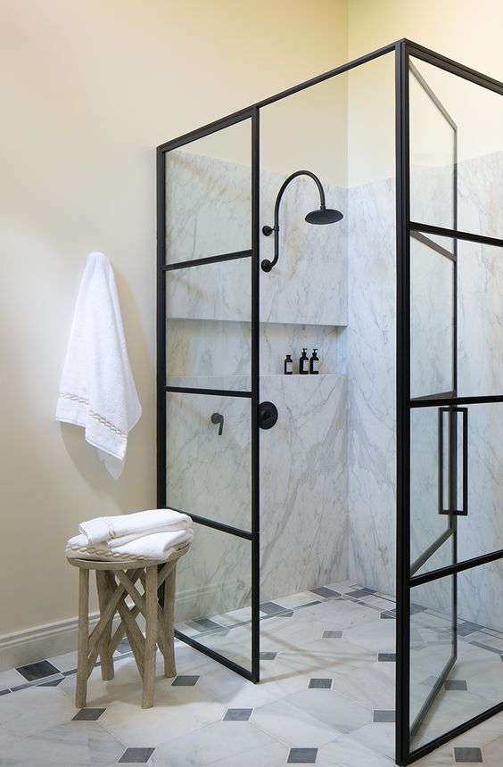 a beautiful neutral bathroom with tan walls, white marble and black and white tiles, a shower space with black frame doors and a stool