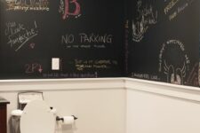 a black and white powder room with black chalkboard walls, white paneling, a wooden vanity and a toilet, some art on the walls