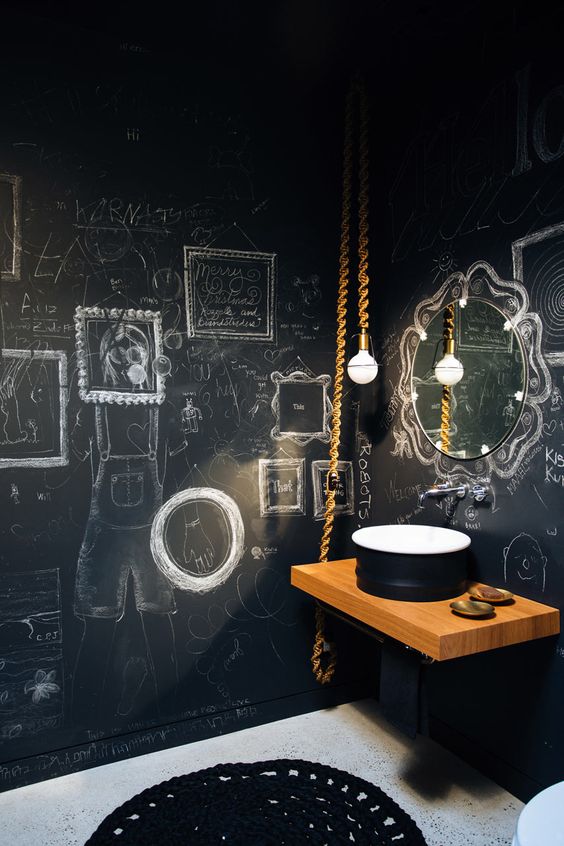 a catchy bathroom with black chalkboard walls, a wooden wall-mounted vanity and a black sink, a bulb on rope