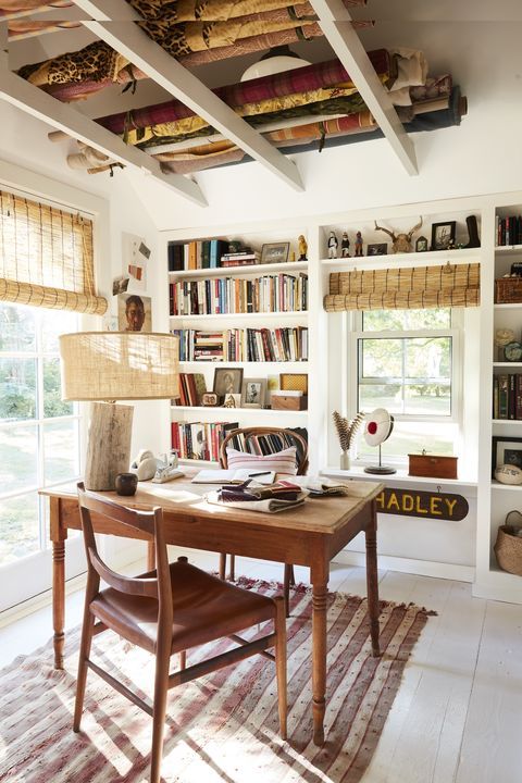 a catchy farmhouse home office with built in storage units and shelves, a wooden desk and chair, a rustic lamp and woven shades