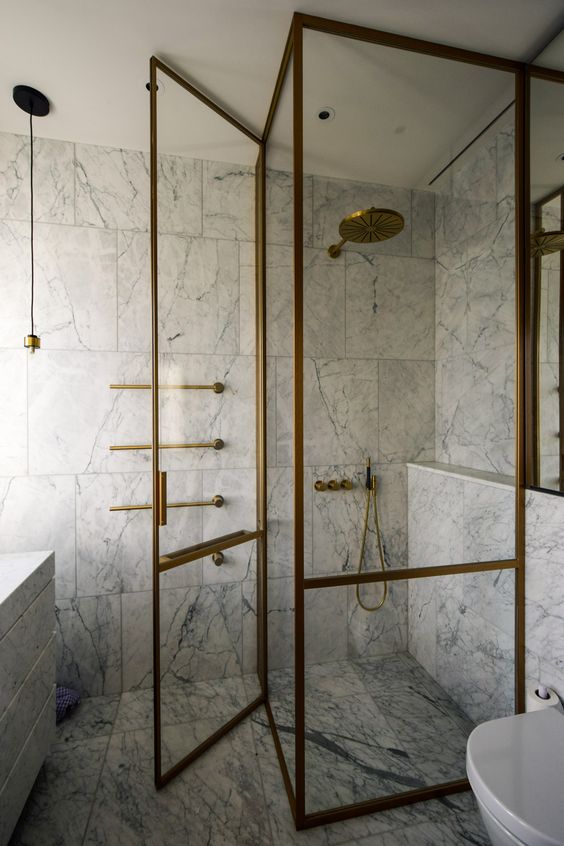 a chic and refined bathroom clad with white marble tiles, with a shower space and with brass frames doors and brass fixtures