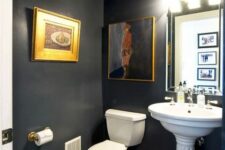 a chic and refined powder room with chalkboard walls, a pedestal sink and a toilet, a mirror with lights and some vintage artwork