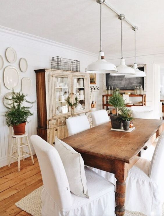 a farmhouse dining area with stained furniture, white covered chairs, potted greenery and white pendant lamps