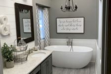 a grey farmhouse bathroom with a large vintage vanity, a stone countertop, an oval tub, a chandelier and some vintage wall lamps