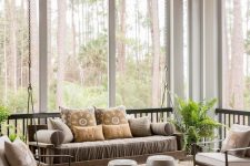 a grey sunroom with a suspended daybed, neutral chairs, round side tables and a printed rug, potted greenery