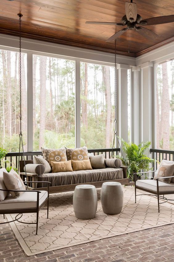 a grey sunroom with a suspended daybed, neutral chairs, round side tables and a printed rug, potted greenery