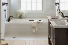 a light-filled farmhouse bathroom with a wooden floor, white tiles, dark wooden vanities and black fixtures