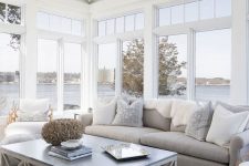 a modern airy sunroom with a grey sofa and neutral pillows, a coffee table and a pouf, some rugs and blankets is welcoming