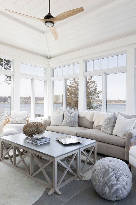 a modern airy sunroom with a grey sofa and neutral pillows, a coffee table and a pouf, some rugs and blankets is welcoming