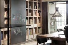 a modern and welcoming home office with a large bookcase, a chalkboard, a large shared desk and black chairs, pendant lamps