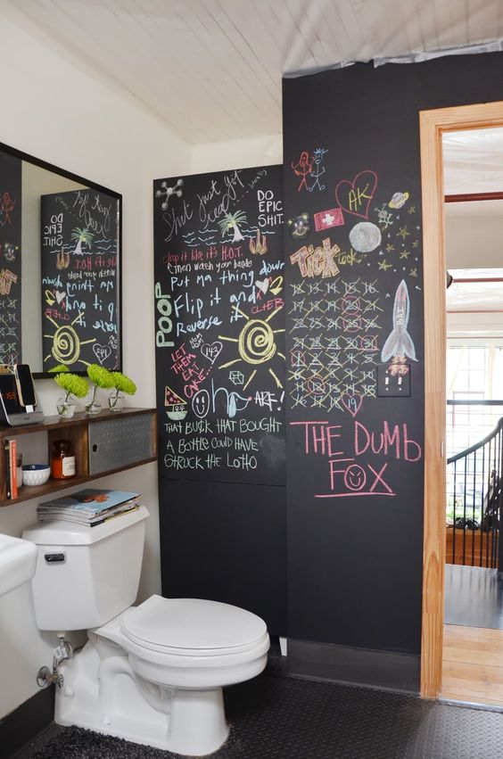 a modern bathroom with a chalkboard accent wall, a black tile floor, bookshelves, a mirror, a white toilet and fun decor on the wall