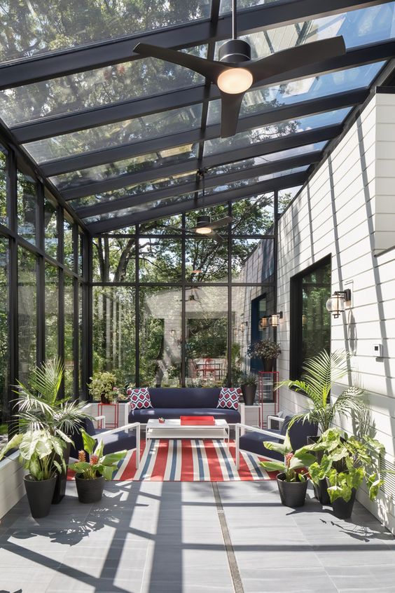 a modern fully glazed sunroom with white furniture and navy upholstery, potted plants and bright red touches here and there