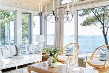 a modern neutral sunroom with a sea view, white seating furniture, rattan chairs, a low coffee table and a vintage chandelier