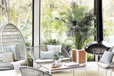 a modern neutral sunroom with lightweight wicker furniture, a suspended chair, a black suspended fireplac,e potted greenery and lots of light