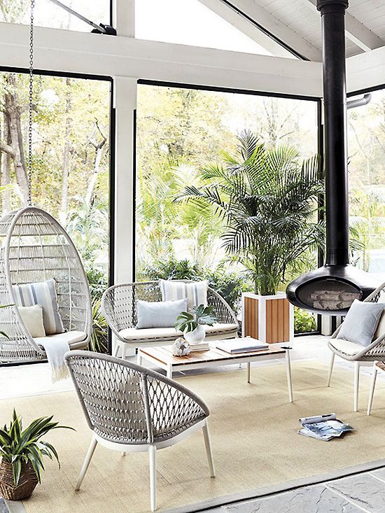 a modern neutral sunroom with lightweight wicker furniture, a suspended chair, a black suspended fireplac,e potted greenery and lots of light