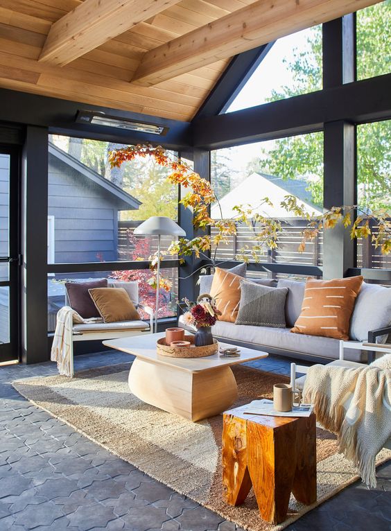 a modern sunroom with a grey sofa, a low coffee table, creamy chairs, a staines stool and fall leaves for decor