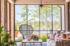 a modern sunroom with glazed walls, a neutral sofa, a coffee table and a peacock chair, a potted plant and cool views