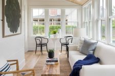 a neutral modern sunroom with a white sofa, some pillows, a white table and black chairs, a wooden coffee table and a woven chair